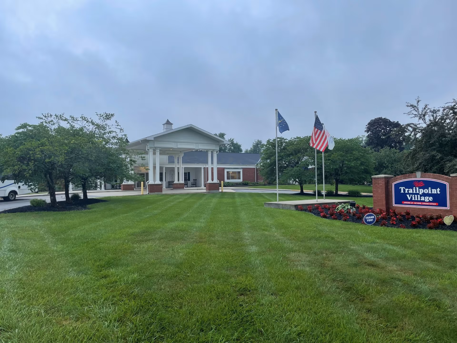 Exterior view of Trailpoint Village senior living facility showing a well-maintained lawn, a covered entrance with white columns, three flagpoles with flags, and a sign with the facility name surrounded by flowers.