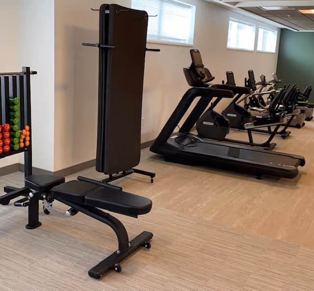 Interior view of a fitness room with exercise equipment including a weight bench, a rack of colorful dumbbells, a folded exercise mat, treadmills, and stationary bikes lined up against a wall with windows.