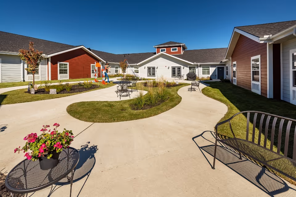Outdoor courtyard area of a senior living facility with paved walkways, green grass, small trees, and flower beds. There are metal benches and a small table with a potted plant. The surrounding buildings have red and white siding with multiple windows under a clear blue sky.