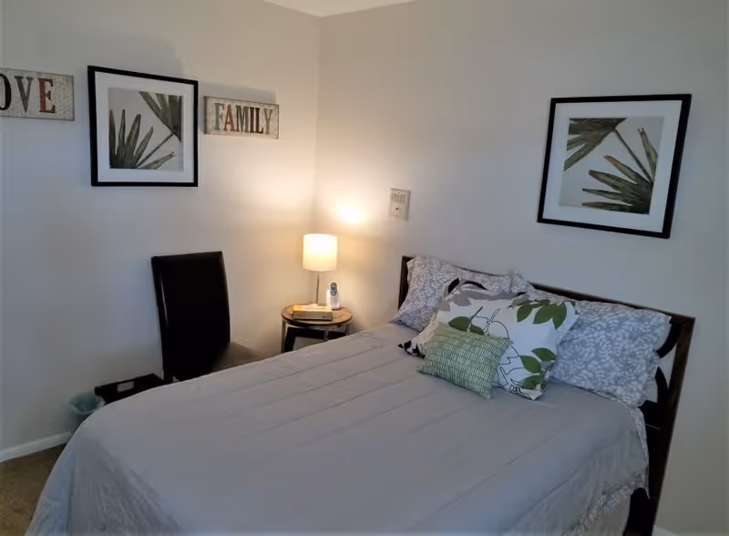 A bedroom with a neatly made bed featuring multiple pillows with leaf patterns. There is a small round bedside table with a lamp and a phone on it. A black chair is placed against the wall. The walls have framed botanical prints and decorative signs that say 'LOVE' and 'FAMILY'.