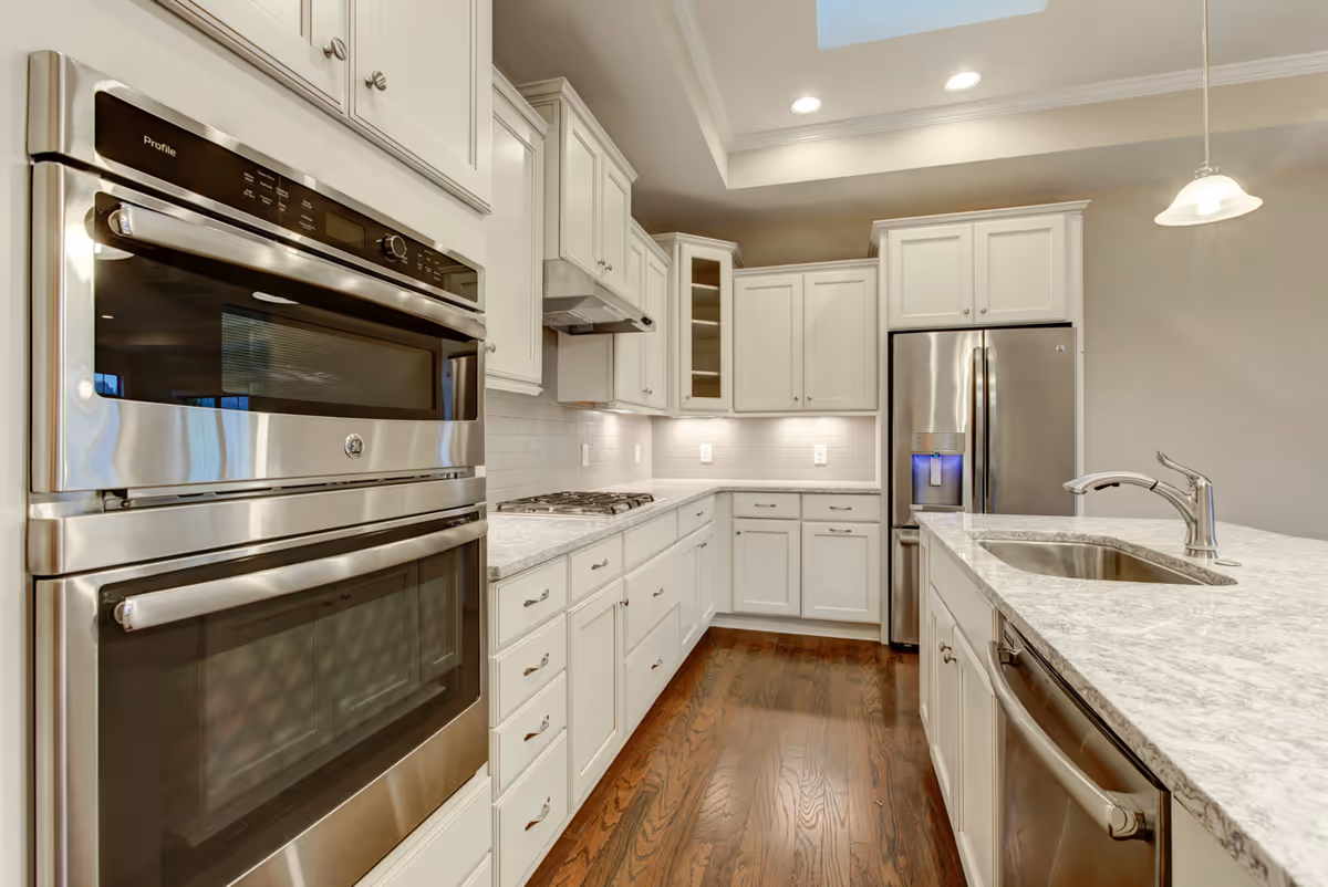 Modern kitchen with white cabinetry, stainless steel double oven, refrigerator, dishwasher, and a marble countertop island with a sink. The kitchen has hardwood floors and recessed lighting with a skylight above.