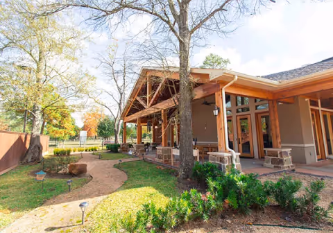 Covered wooden porch and patio of a senior living building with a winding gravel path and landscaped yard.