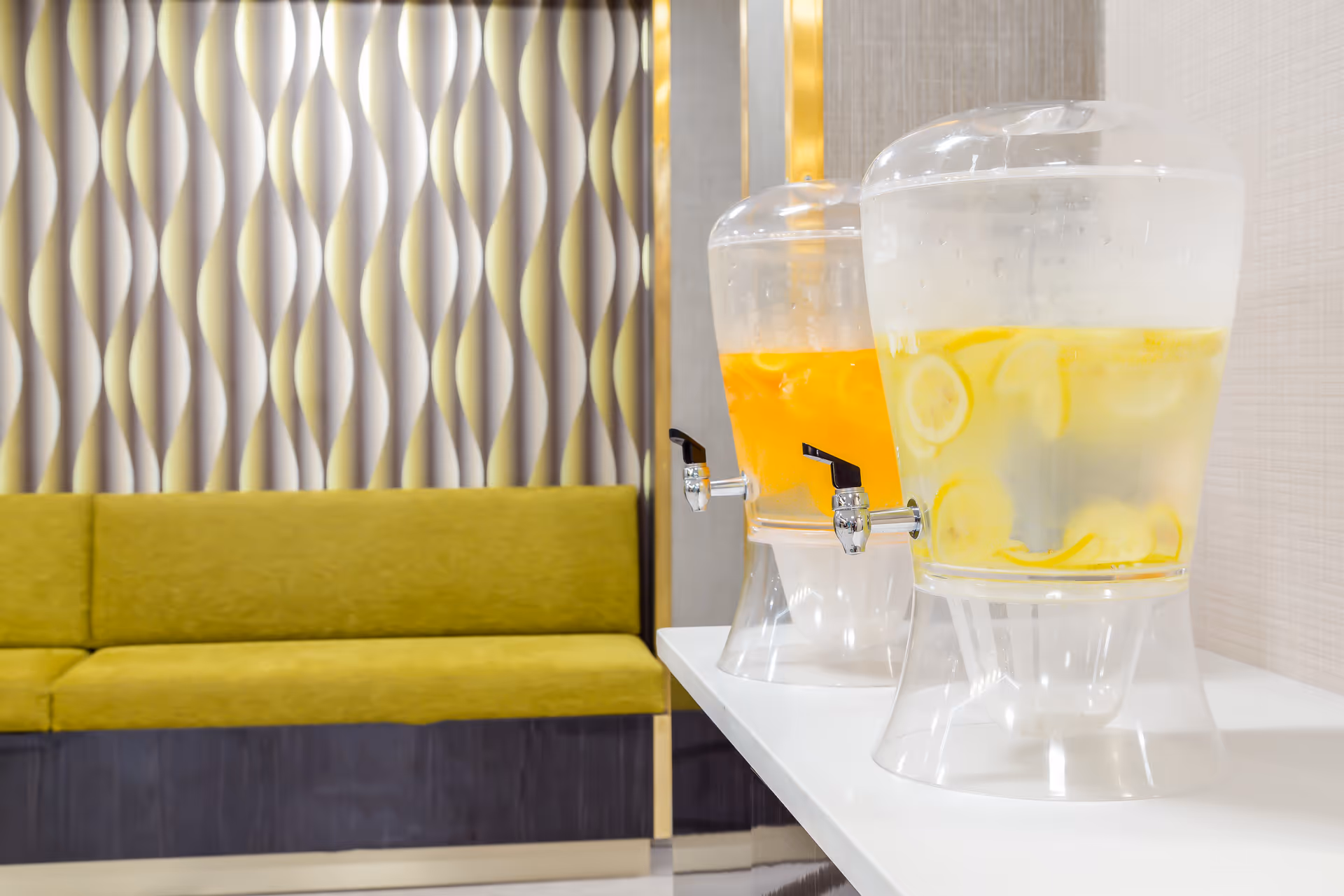 Two large beverage dispensers on a white counter, one filled with lemon water and the other with an orange-colored drink, next to a green cushioned bench and a wall with a decorative wavy pattern.