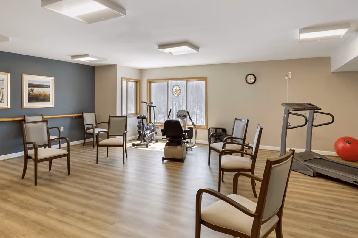 A senior living facility exercise room with several wooden chairs arranged in a circle on a wood floor. The room has large windows letting in natural light, exercise equipment including a treadmill, stationary bike, and elliptical machine, and a red exercise ball. The walls are painted beige and blue with two framed pictures and a clock.