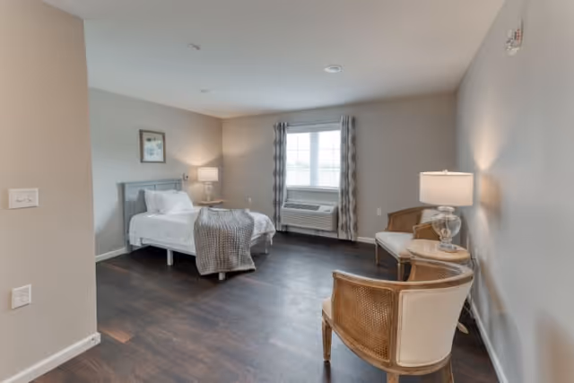 A spacious senior living bedroom with a single bed made with white linens and a gray throw blanket. The room features dark wood flooring, a window with patterned curtains, an air conditioning unit below the window, two wooden chairs with light cushions, a small round table with a lamp, and neutral-colored walls.