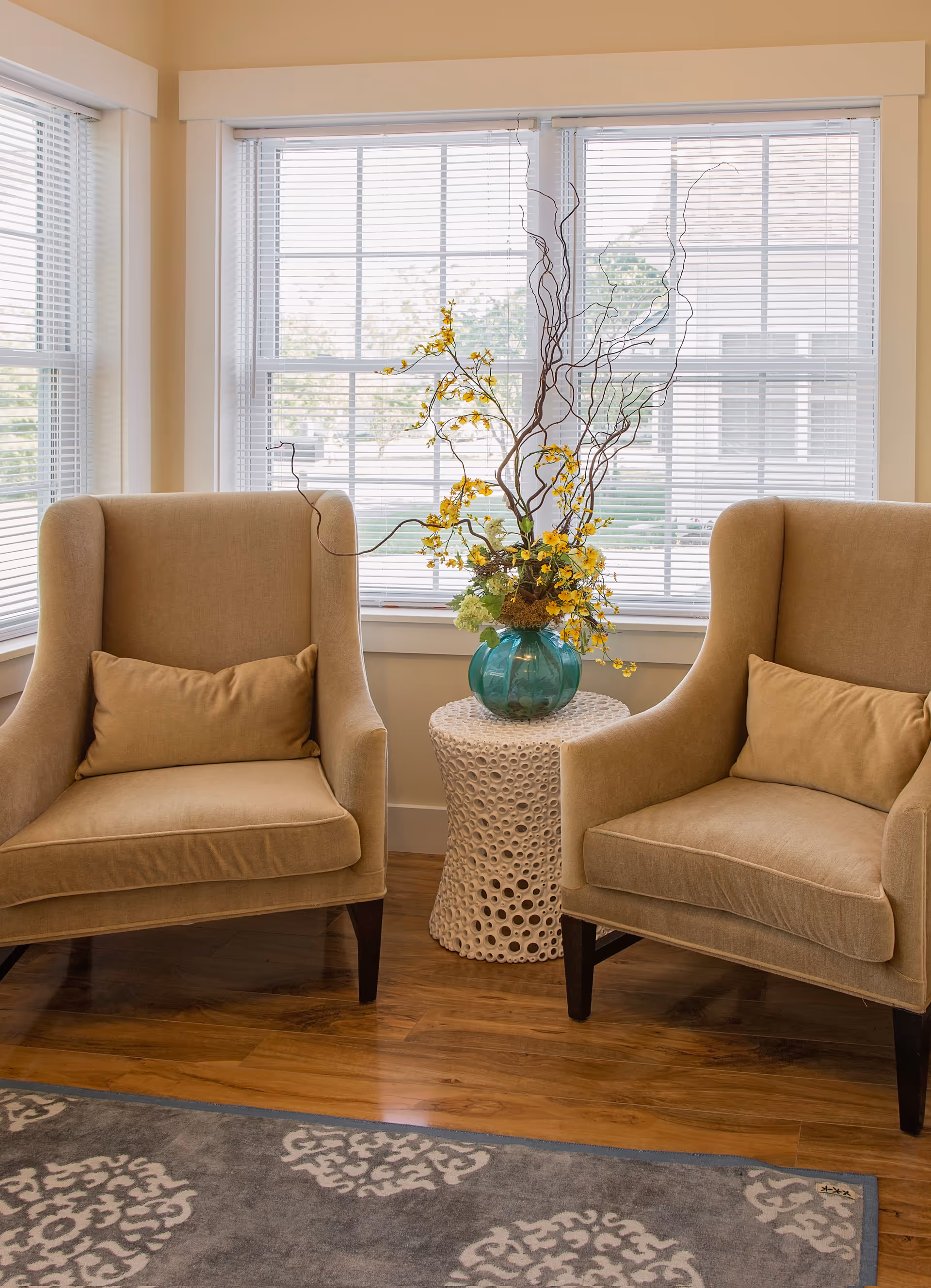 Two beige armchairs flank a small white side table holding a turquoise vase with yellow flowers in front of a sunlit window.