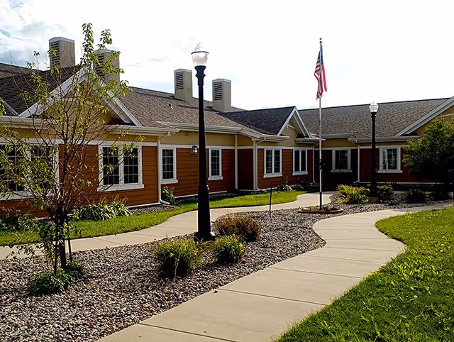 Exterior view of a single-story building with brown and beige siding, multiple windows, and a shingled roof. A curved concrete walkway leads to the entrance, flanked by landscaped areas with rocks, small bushes, and young trees. Two black lamp posts and an American flag on a flagpole are visible along the walkway under a partly cloudy sky.