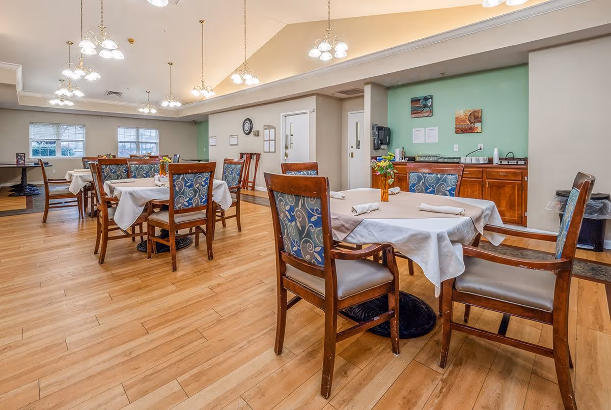 A dining room in a senior living facility with several wooden tables covered with white tablecloths and set with rolled napkins. Each table is surrounded by wooden chairs with blue patterned cushions. The room has wooden flooring, multiple ceiling light fixtures, and a green accent wall with cabinets and a countertop in the background.
