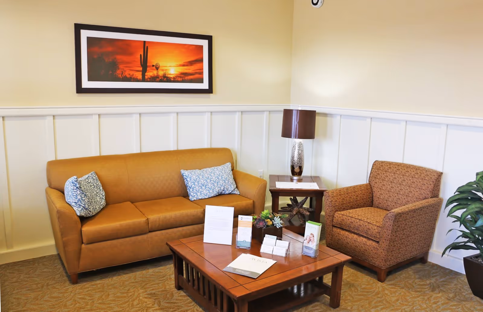 A cozy seating area with a brown sofa adorned with two patterned blue pillows, a brown patterned armchair, a wooden coffee table with brochures and decorative items, a side table with a lamp, and a framed picture of a desert sunset with a cactus on the wall.
