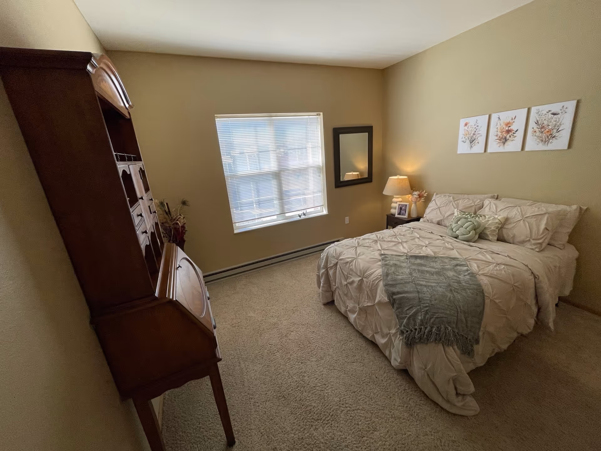 A cozy bedroom with a neatly made bed featuring cream-colored bedding and a green throw blanket. Above the bed are three floral paintings. To the right of the bed is a nightstand with a lamp, a framed photo, and a small flower arrangement. A window with blinds lets in natural light, and a wooden cabinet with shelves stands against the left wall. The walls are painted beige and the floor is carpeted.