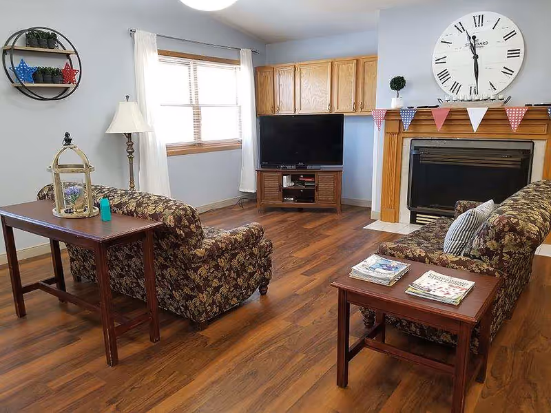 Cozy living room with floral sofas, wooden side tables, a TV stand, fireplace with a large wall clock, and hardwood floors.
