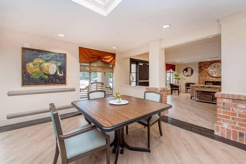 A bright and clean interior space featuring a wooden table with four chairs around it. The room has light-colored walls and flooring, with a painting of pears on one wall. There is a window with a red valance and a view of the outside. In the background, there is a seating area with chairs, a large clock on a brick wall, and a round mirror.