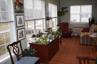 Bright communal sitting room with large windows, tiled floor, upholstered chairs and a sofa, and a central table covered with potted plants.