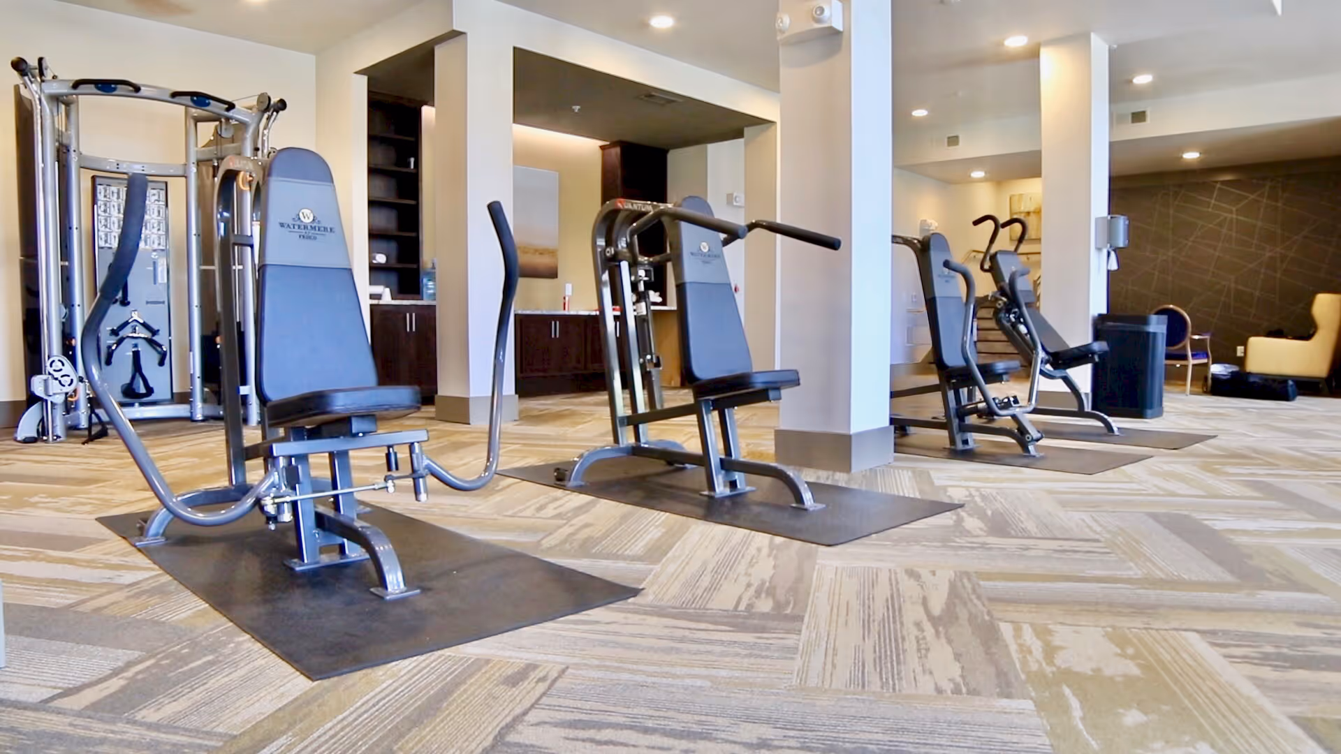 A fitness room with multiple exercise machines including seated weight machines and elliptical trainers, set on a patterned carpeted floor. The room has neutral-colored walls, recessed lighting, and a seating area with chairs in the background. The exercise machines have the Watermere at Frisco logo on their backrests.
