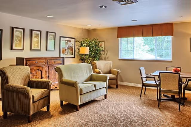 A cozy living room area with two armchairs and a loveseat arranged around a wooden sideboard with framed artwork above it. There is a potted plant and a floor lamp in the corner near a window with blinds and an orange valance. A small dining table with four chairs is positioned near the window.