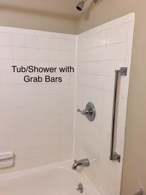 A bathtub and shower area with white tiled walls, a metal grab bar mounted vertically on the right wall, a showerhead at the top, and a faucet with controls below. The text 'Tub/Shower with Grab Bars' is displayed on the tiles.