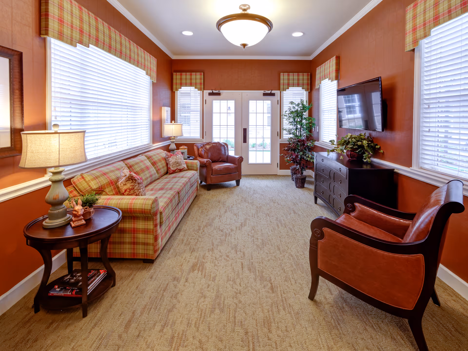 A cozy living room with orange walls and beige carpet. The room features a plaid sofa with matching window valances, two brown leather armchairs, a dark wooden side table with a lamp and small decorations, a dark wooden dresser with plants on top, and a wall-mounted flat-screen TV. Large windows with white blinds let in natural light, and double glass doors are at the far end of the room.