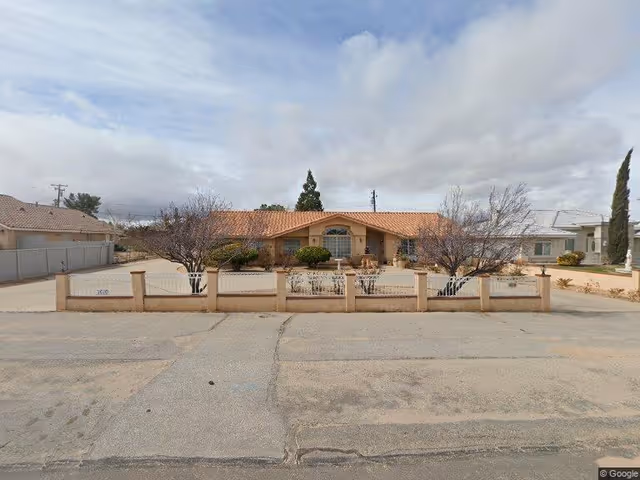 Single-story stucco building with a tile roof, low front wall and driveway, and sparse desert landscaping under a cloudy sky.