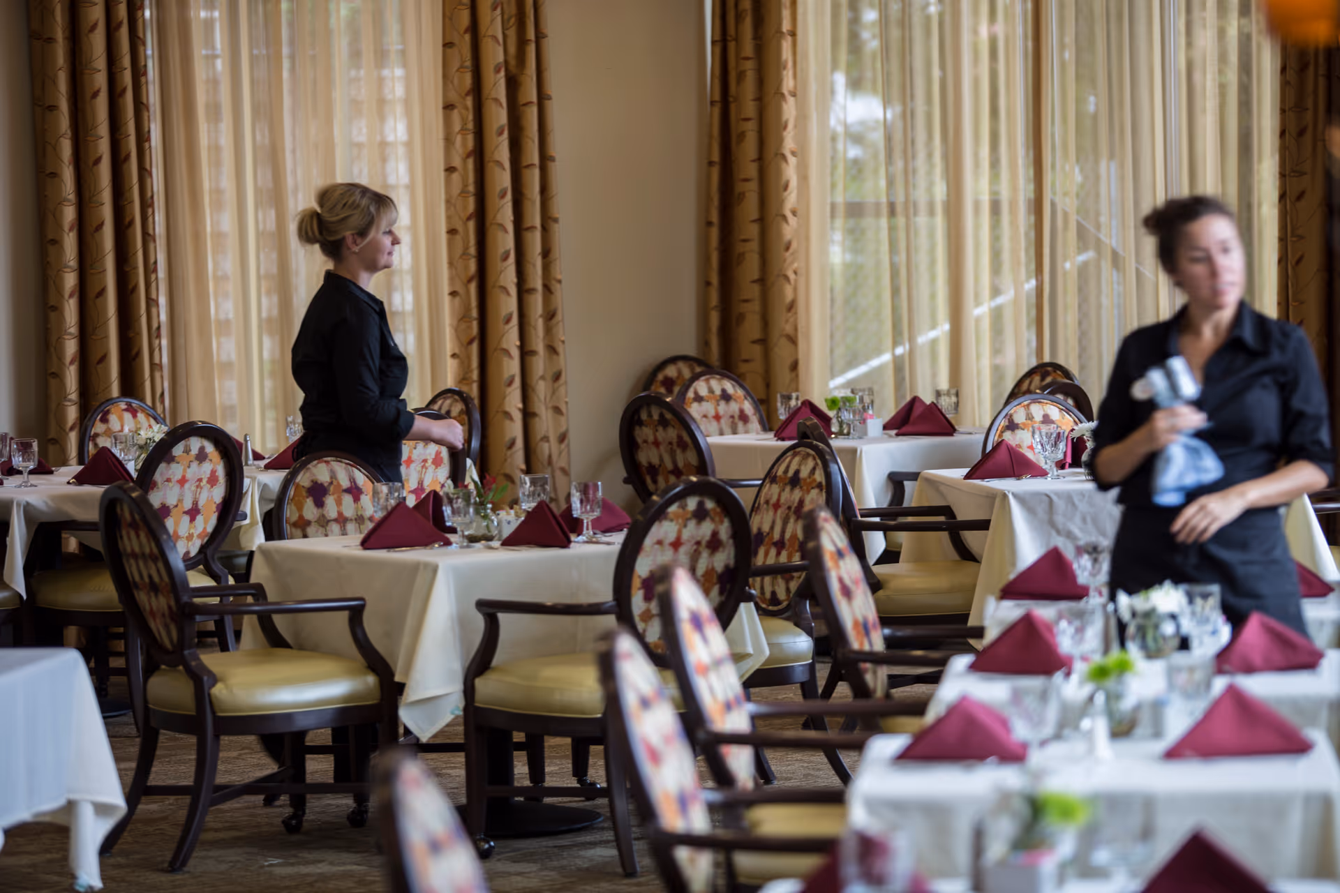 Dining room with tables covered in white tablecloths and set with folded burgundy napkins, glassware, and small floral centerpieces. Two women dressed in black are present, one standing near a table and the other holding a cloth, with large windows and patterned curtains in the background.