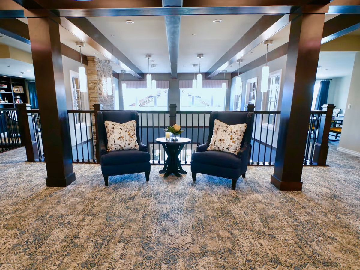 A cozy seating area with two dark blue armchairs featuring floral patterned pillows, separated by a small round table with a flower arrangement and a candle. The area has a patterned carpet, dark wooden beams on the ceiling, and pendant lights hanging above. Large windows in the background allow natural light to fill the space.