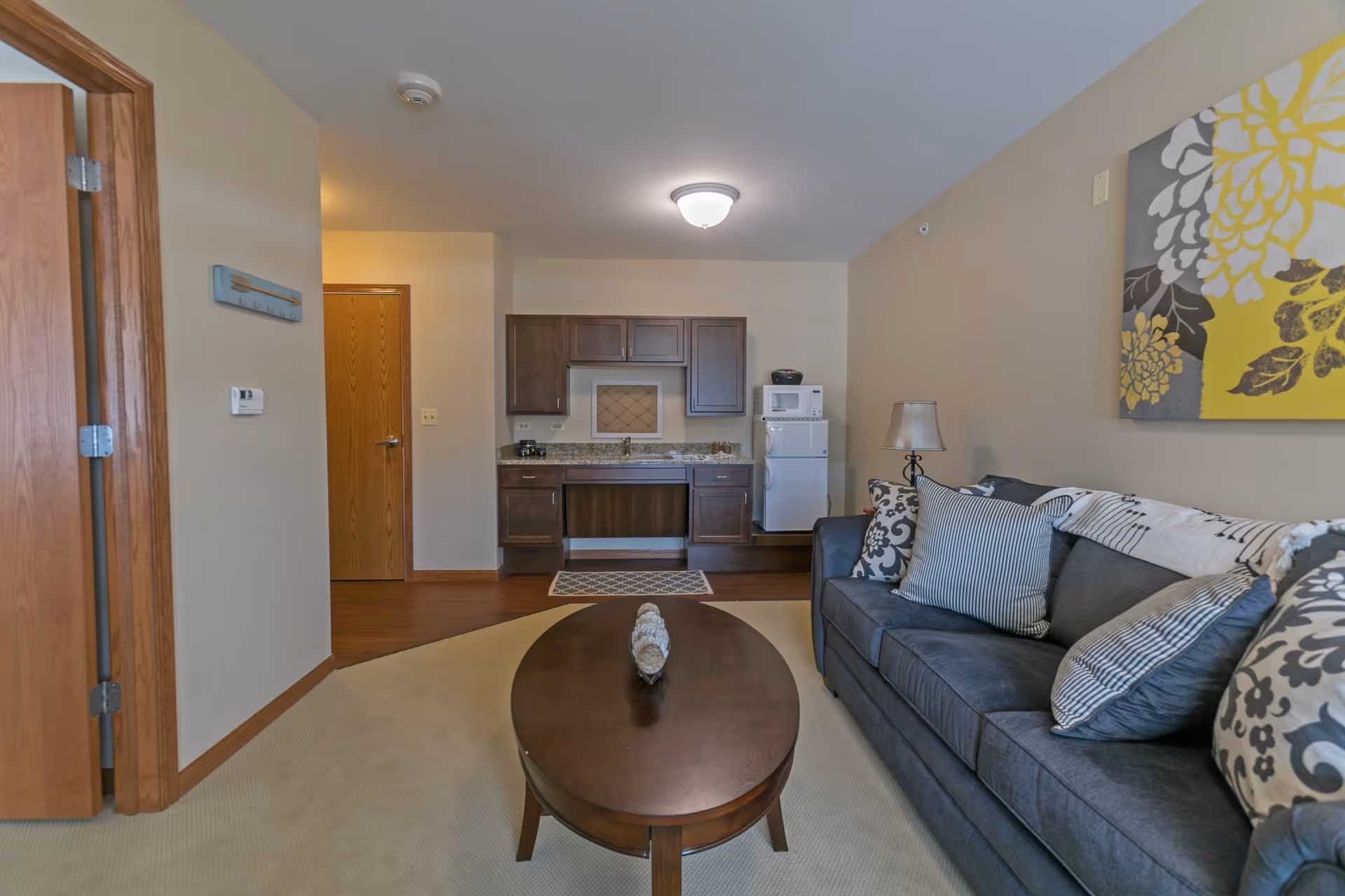 A cozy living area in a senior living facility featuring a dark gray sofa with multiple patterned pillows, a wooden oval coffee table with a decorative centerpiece, a small kitchenette with dark wood cabinets, a white refrigerator, microwave, and a lamp on a side table. The walls are beige with a large floral artwork in yellow, gray, and white hanging above the sofa. There are wooden doors and a carpeted floor transitioning to hardwood near the kitchenette.