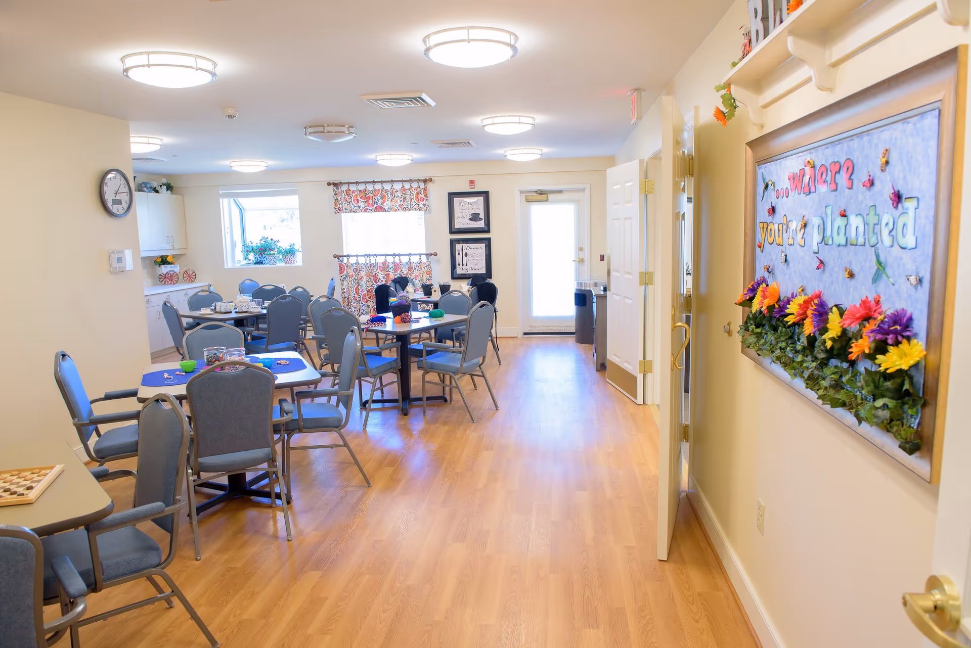 Bright communal dining/activity room with multiple round tables and chairs, wood floors, and a colorful wall display.