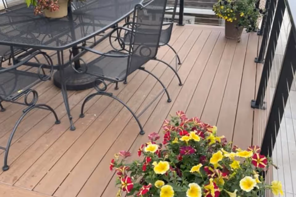 A wooden outdoor deck with metal patio table and chairs and colorful potted flowers by a railing.