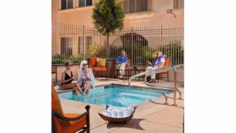 Four older adults sitting and relaxing around a small outdoor hot tub/pool on a sunny patio with lounge chairs.