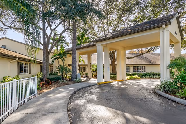 Covered porte-cochère entrance and circular driveway of a single-story building surrounded by trees and landscaping.