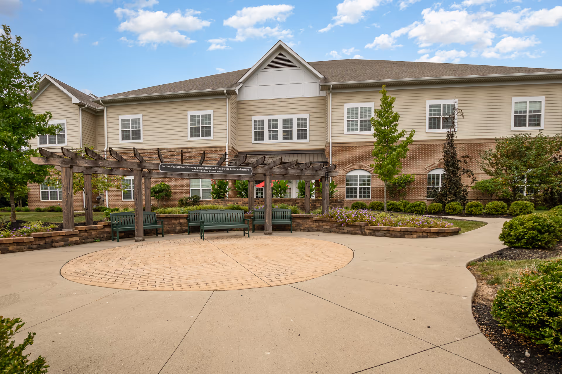 Outdoor garden area with a wooden pergola and green benches arranged in a semicircle. The garden is surrounded by a paved walkway, flower beds, and shrubs. In the background, there is a two-story building with beige siding and brick accents under a partly cloudy sky.