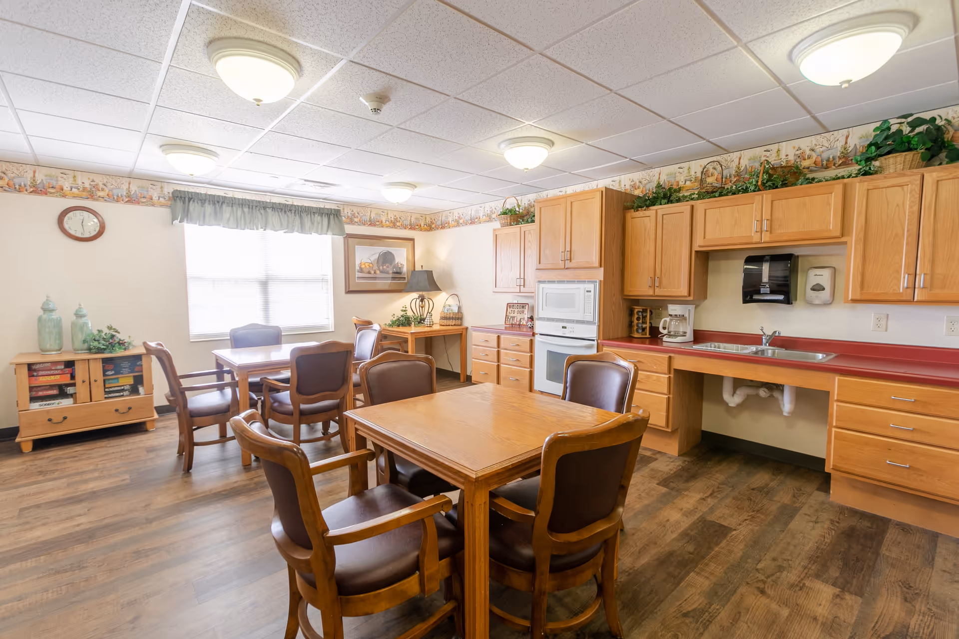Bright communal dining area with wooden tables and chairs and a kitchenette with cabinets, sink, and microwave.
