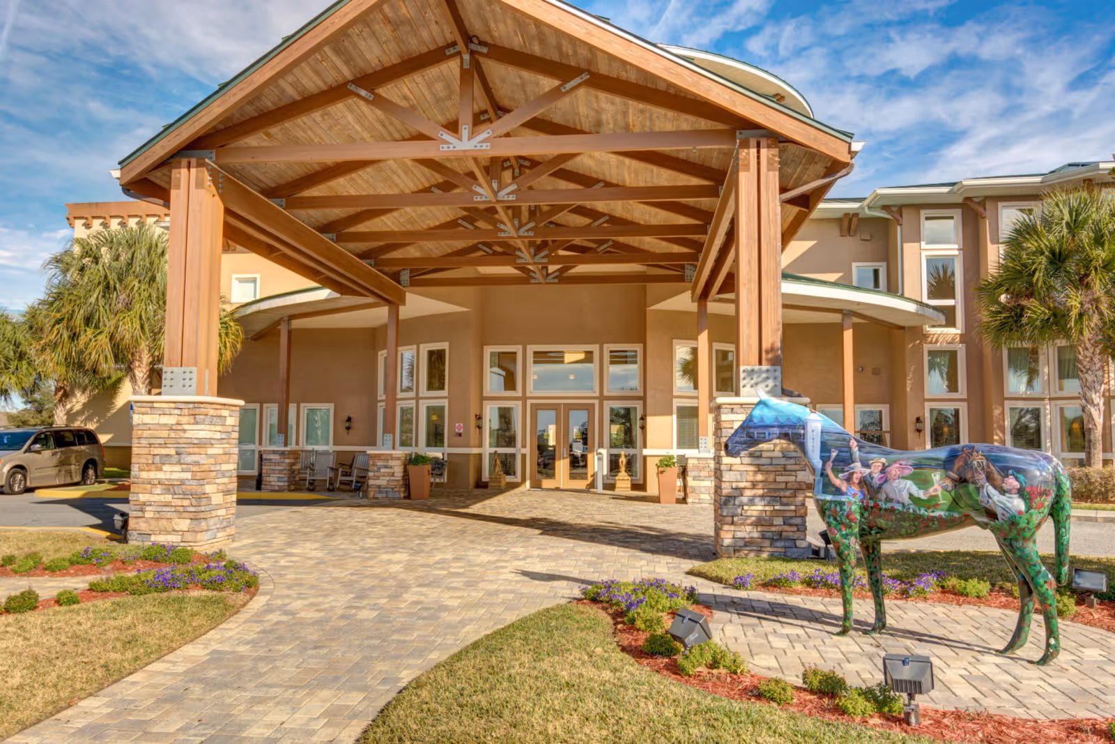 Entrance of a senior living facility with a large wooden canopy supported by stone pillars. There is a colorful horse statue with painted images of people and a horse on it near the entrance. The building has multiple windows and palm trees around the area under a partly cloudy sky.