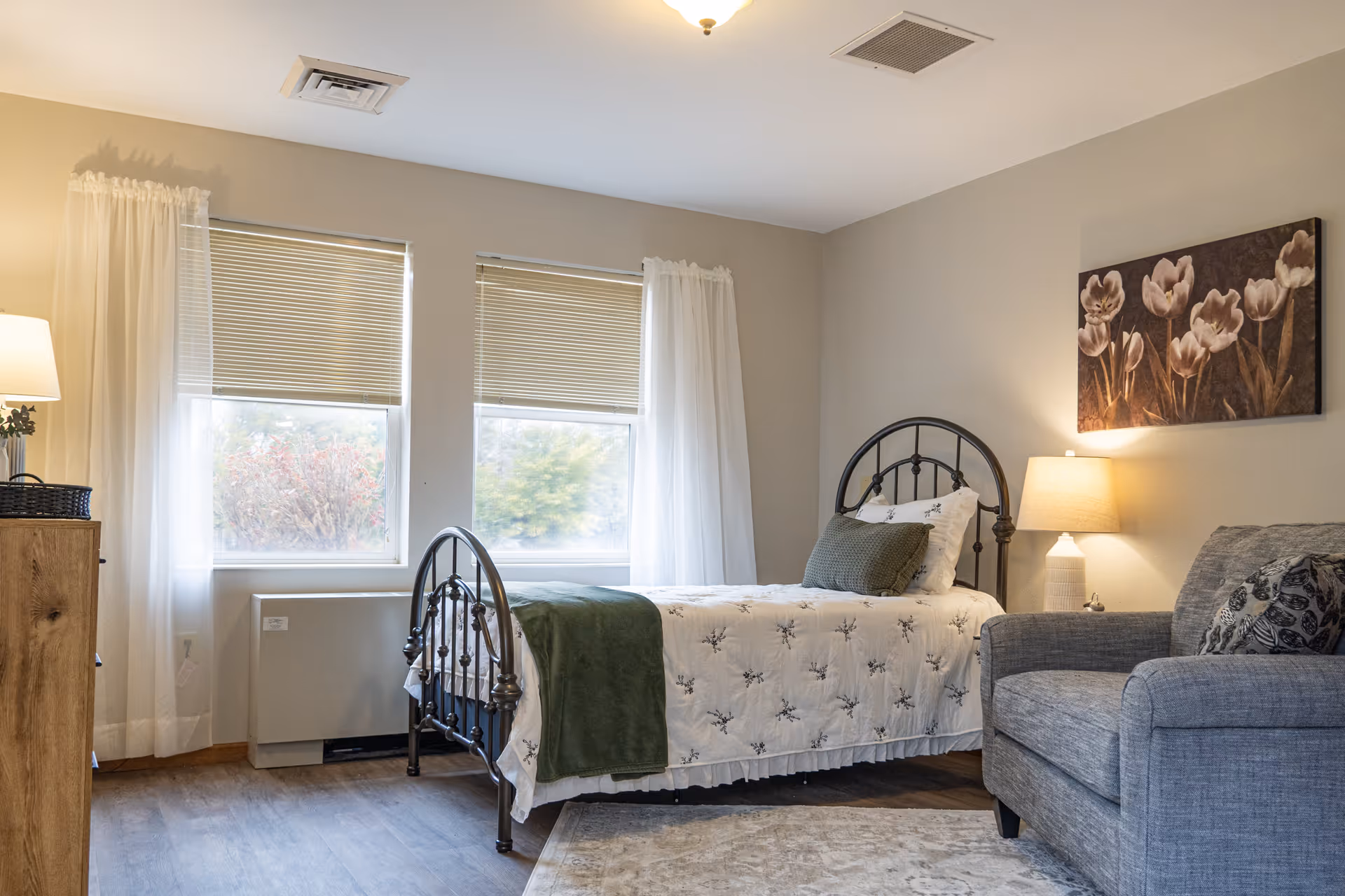 A cozy bedroom with a single metal-framed bed dressed in white floral bedding and a green throw blanket. Next to the bed is a gray upholstered armchair with a patterned pillow. A wooden dresser with a lamp and a basket is on the left side near two windows with blinds and sheer white curtains. A floral painting hangs on the wall above a bedside lamp on a small table.