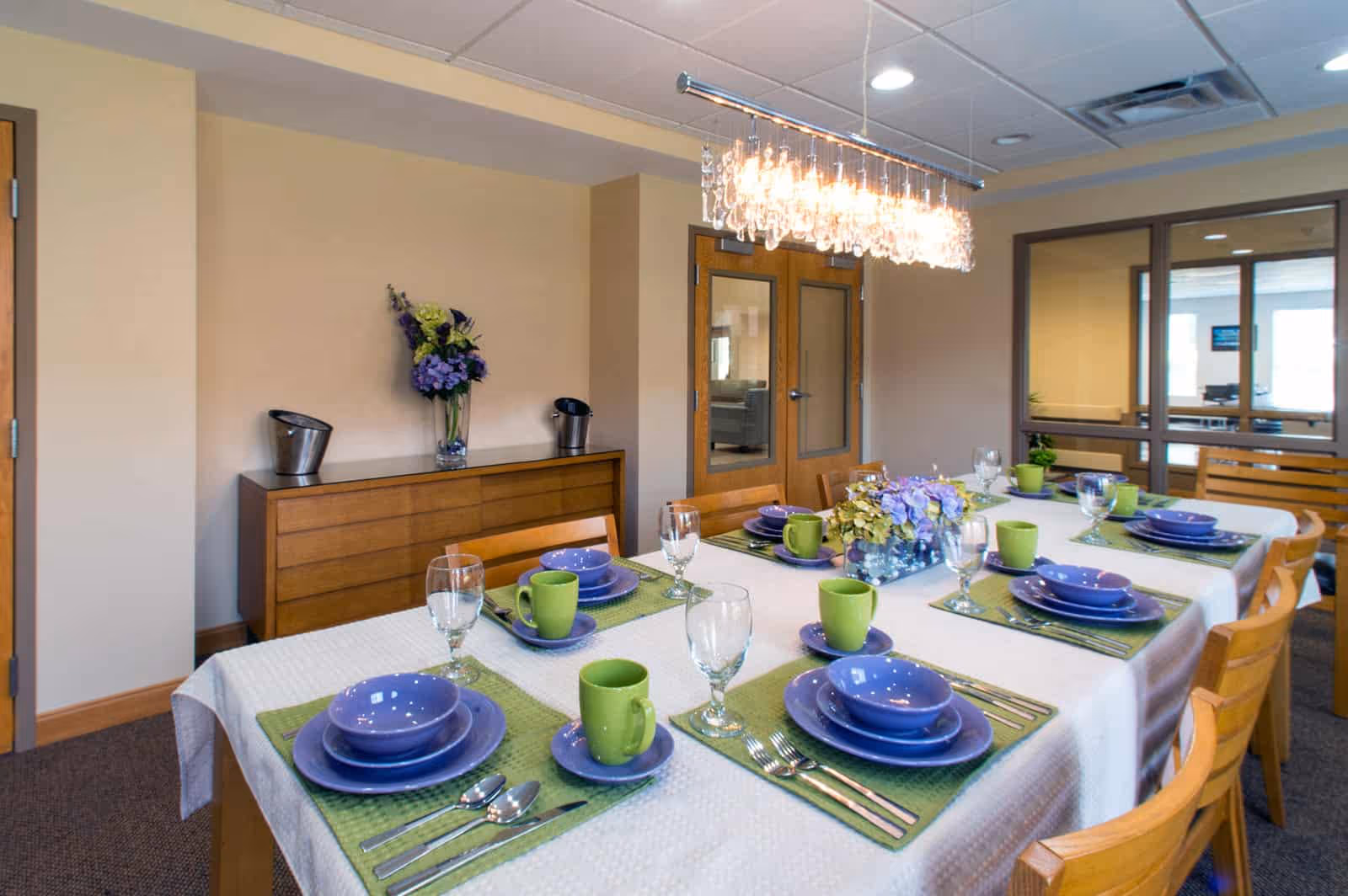 A dining room with a table set for eight people. The table is covered with a white tablecloth and green placemats, each setting includes purple plates, bowls, and green mugs, along with silverware and glasses. A modern chandelier with hanging crystals is above the table. In the background, there is a wooden sideboard with a vase of flowers and two metal ice buckets, and double doors with glass panels. Large windows and a glass partition are also visible.