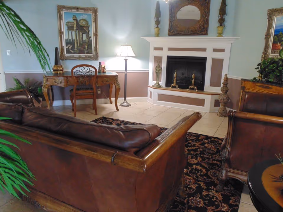 Cozy sitting area with leather sofas facing a fireplace, an ornate desk and lamp, framed artwork and plants.