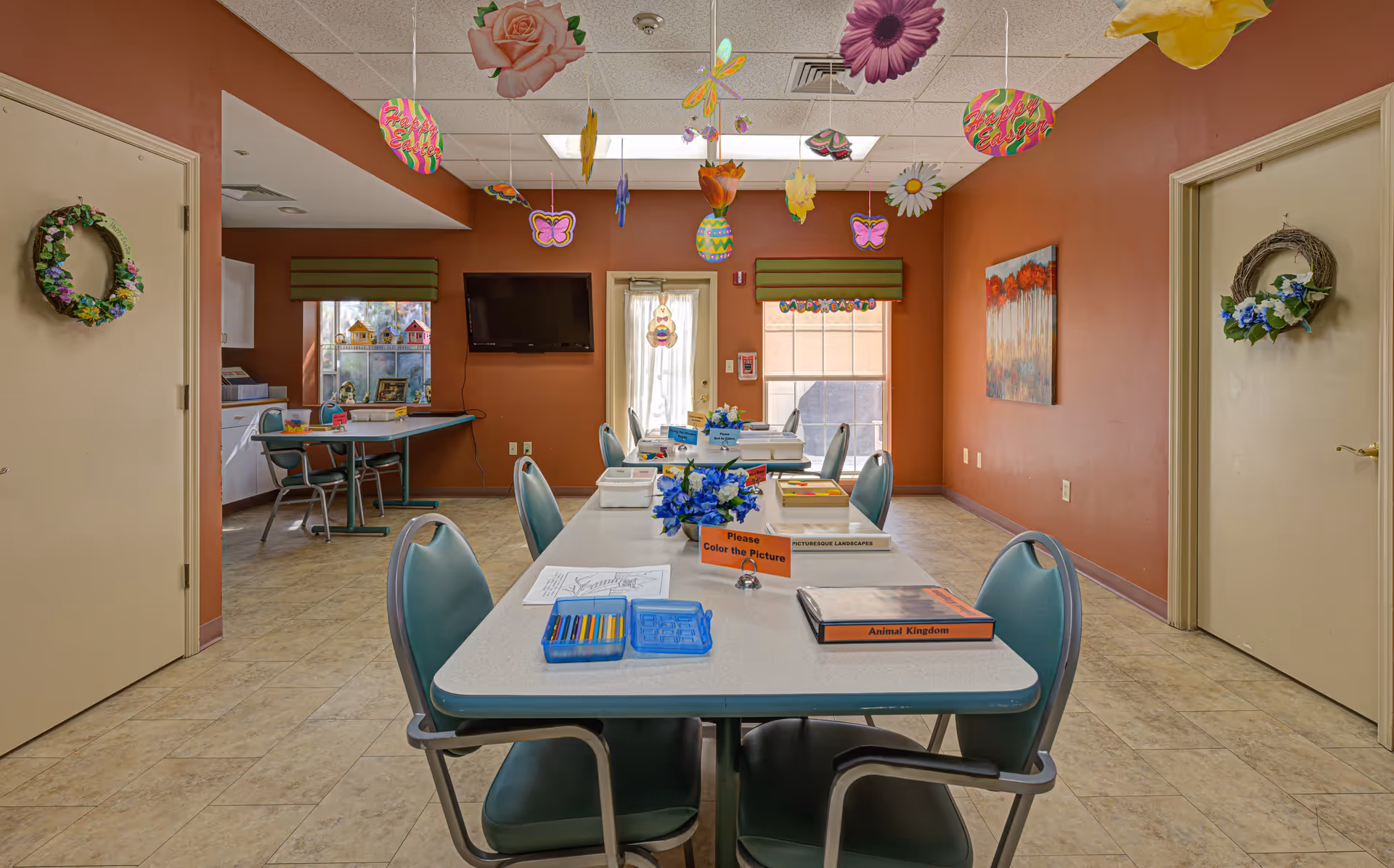 Community activity room with long tables and chairs, hanging floral decorations, and craft supplies arranged for residents.