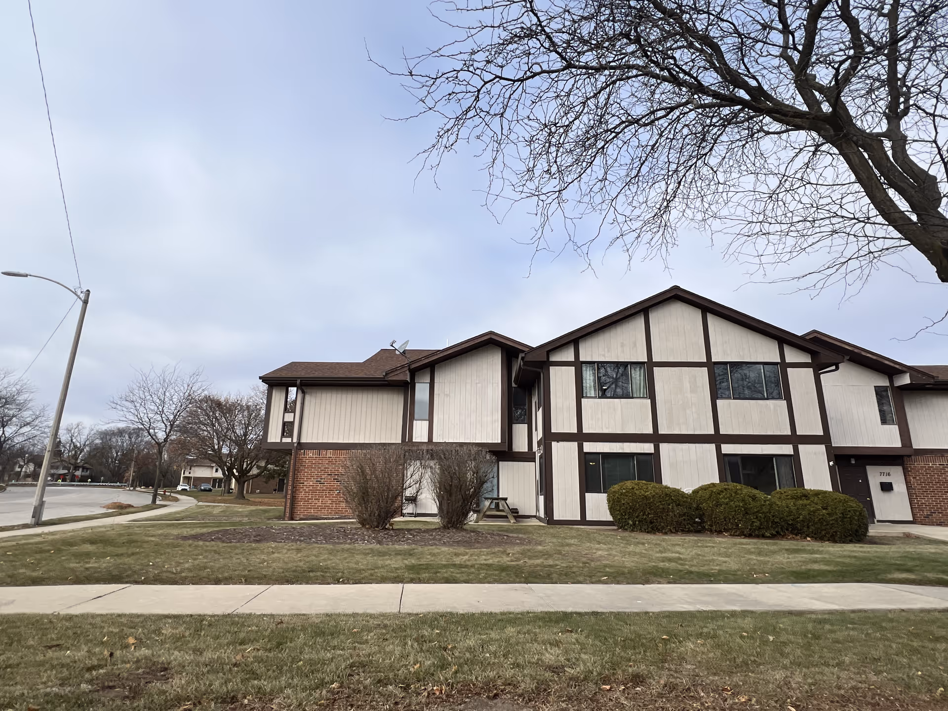 Exterior view of a two-story residential building with beige siding and brown trim, surrounded by grass, bushes, and leafless trees under a cloudy sky.