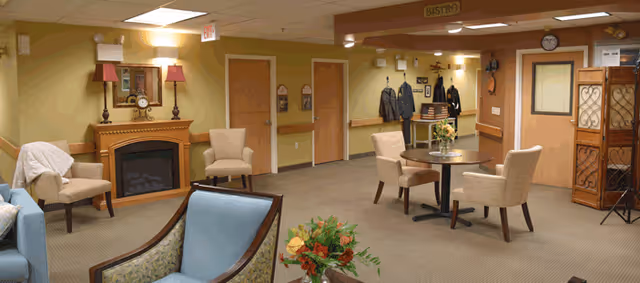 A cozy common area with several upholstered chairs arranged around small tables and a fireplace with a mirror and lamps on the mantel. The walls are painted in warm tones, and there is a coat rack with jackets hanging near the back wall. A folding screen and a clock are also visible.