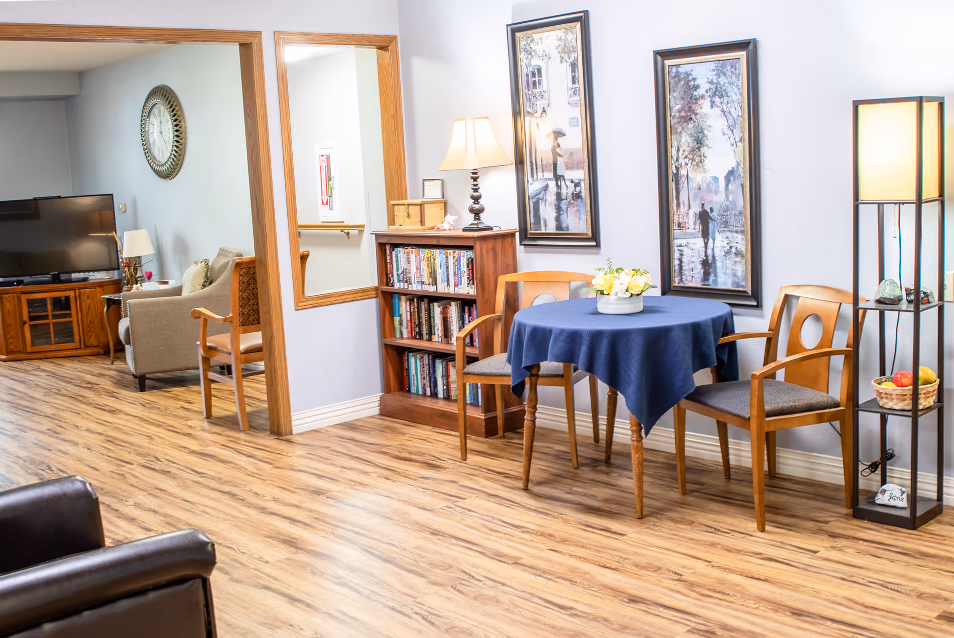 Communal senior living room with a small round table covered by a blue tablecloth, wooden chairs, a bookshelf, framed artwork, and a TV seating area.