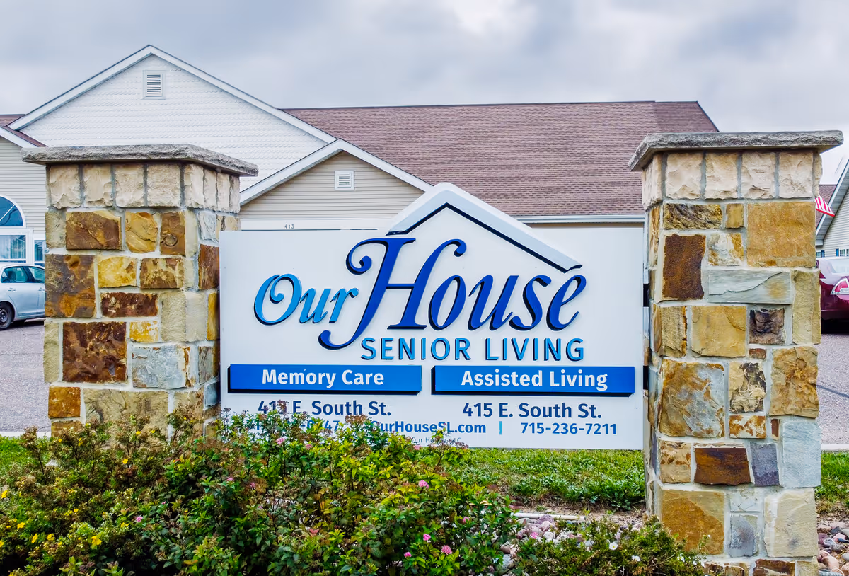 Outdoor sign for Our House Senior Living showing services for Memory Care and Assisted Living, flanked by two stone pillars with a building and parked cars in the background.