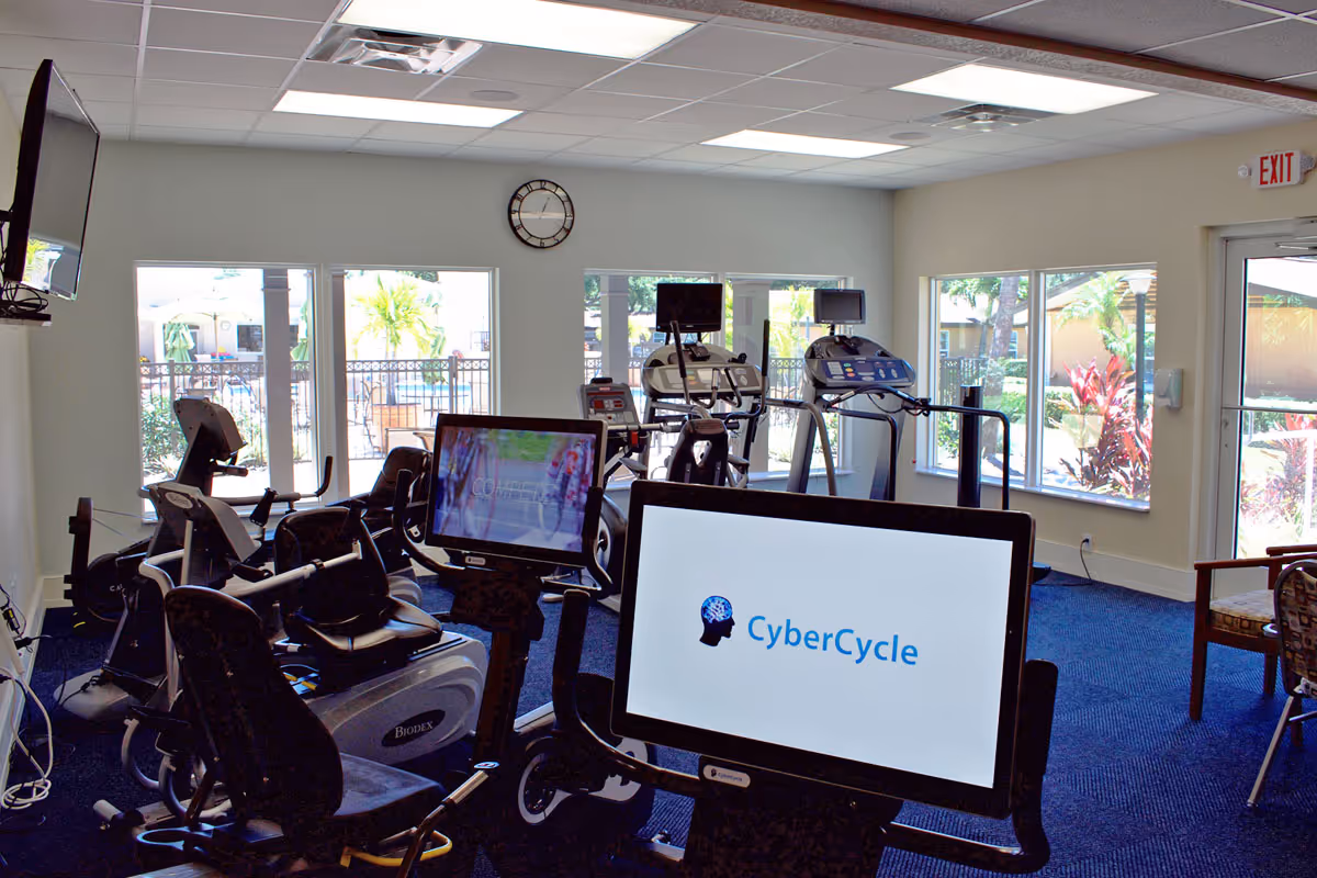 Interior view of a fitness room with exercise equipment including stationary bikes and treadmills, several screens attached to the machines, large windows showing outdoor greenery, a wall clock, and an exit door.