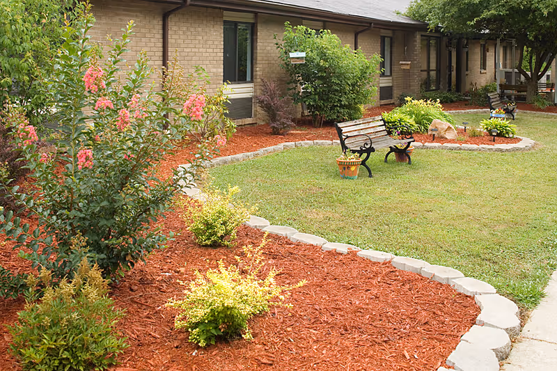 Outdoor garden area at Teays Valley Center featuring a well-maintained lawn, flower beds with red mulch, various shrubs and flowering plants, two wooden benches with decorative flower pots, and a brick building in the background.