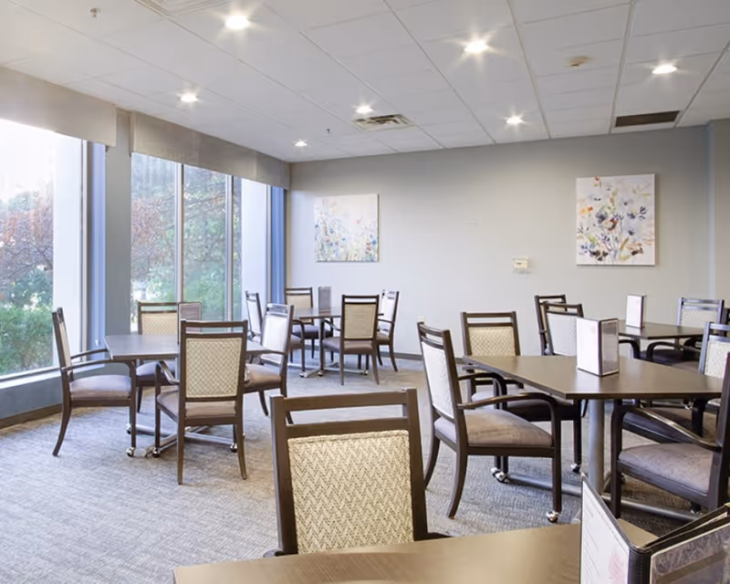 A bright dining room with several tables and chairs arranged neatly. Large windows on one side let in natural light and offer a view of trees outside. The walls are decorated with two floral paintings, and the ceiling has recessed lighting.