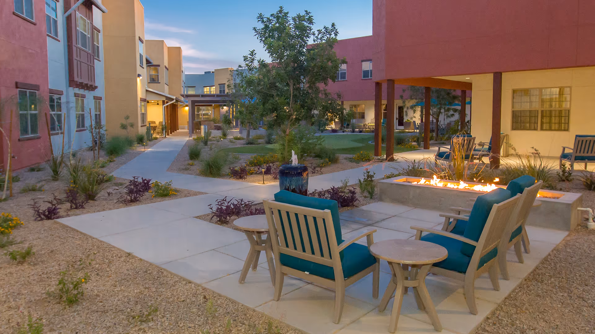Outdoor courtyard area at The Ranch Estates of Tucson featuring a fire pit surrounded by cushioned chairs and small tables, with pathways, plants, and multi-story buildings in the background during dusk.