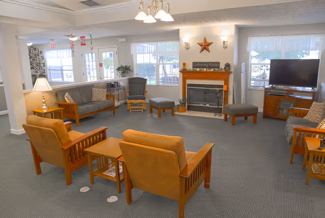 A cozy living room area in a senior living facility with multiple wooden armchairs and sofas arranged around a central coffee table. There is a fireplace with a star decoration above it and a sign that reads 'Gathering Room'. A flat-screen TV is placed on a wooden cabinet to the right. Large windows allow natural light to fill the room.