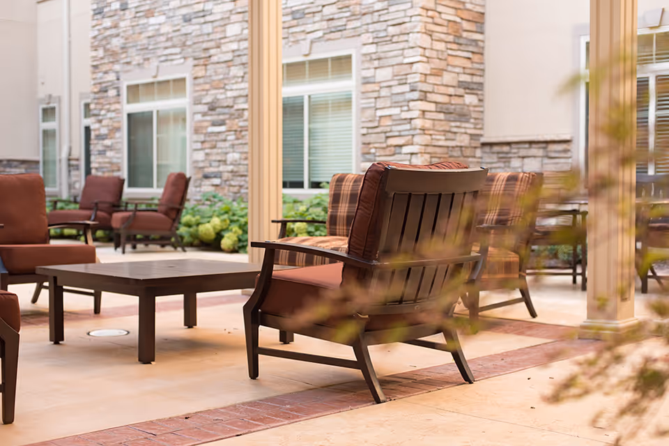 Outdoor patio area with cushioned wooden chairs and a low wooden table, set against a backdrop of a stone and stucco building with windows and some greenery.