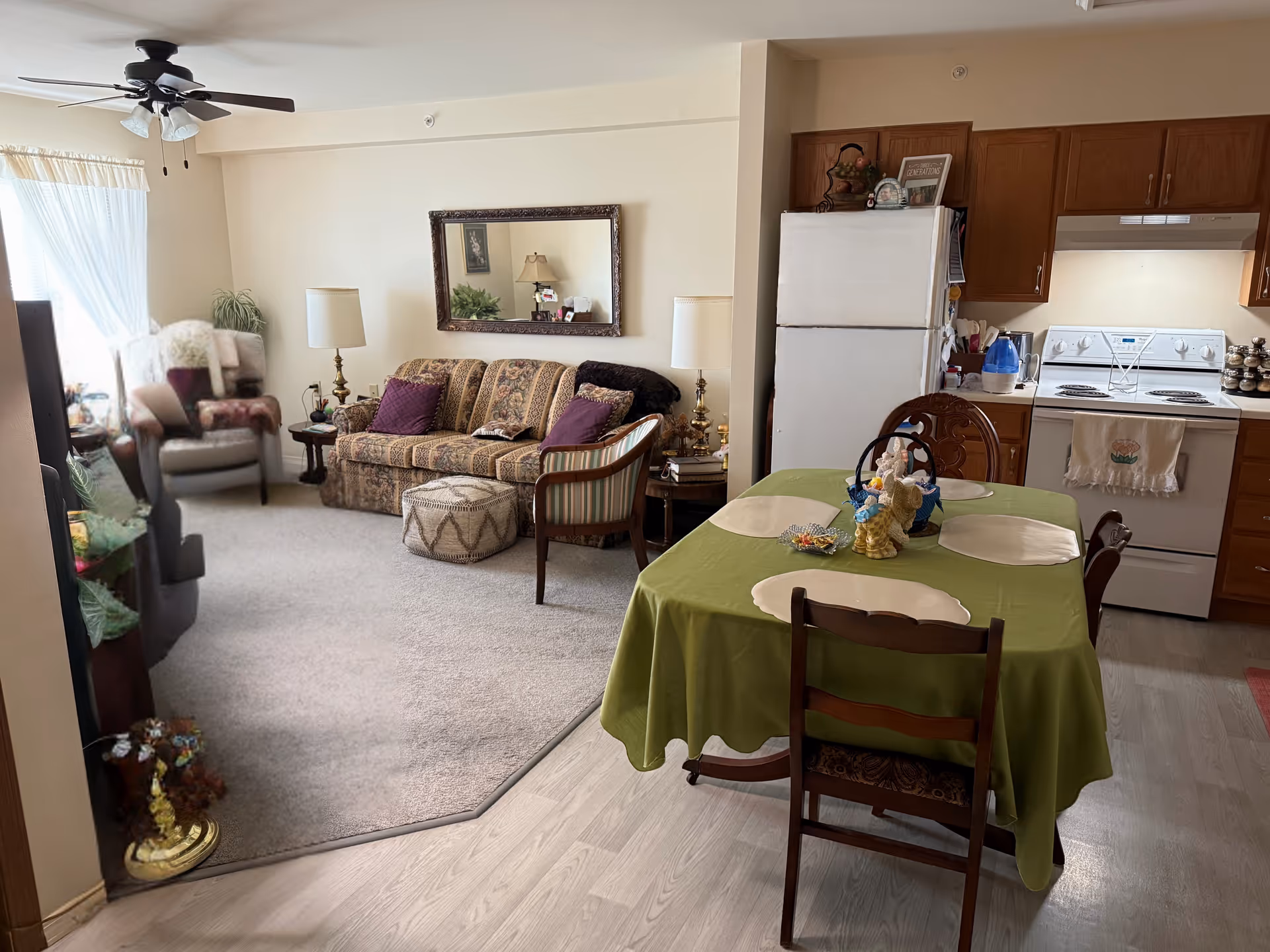 A cozy living and dining area in a senior living facility. The living room has a patterned sofa with purple cushions, an armchair, a small side table with a lamp, and a large mirror on the wall. The dining area features a table covered with a green tablecloth, four chairs, and decorative items on the table. The kitchen area includes a white refrigerator, stove, wooden cabinets, and a countertop with various kitchen items.
