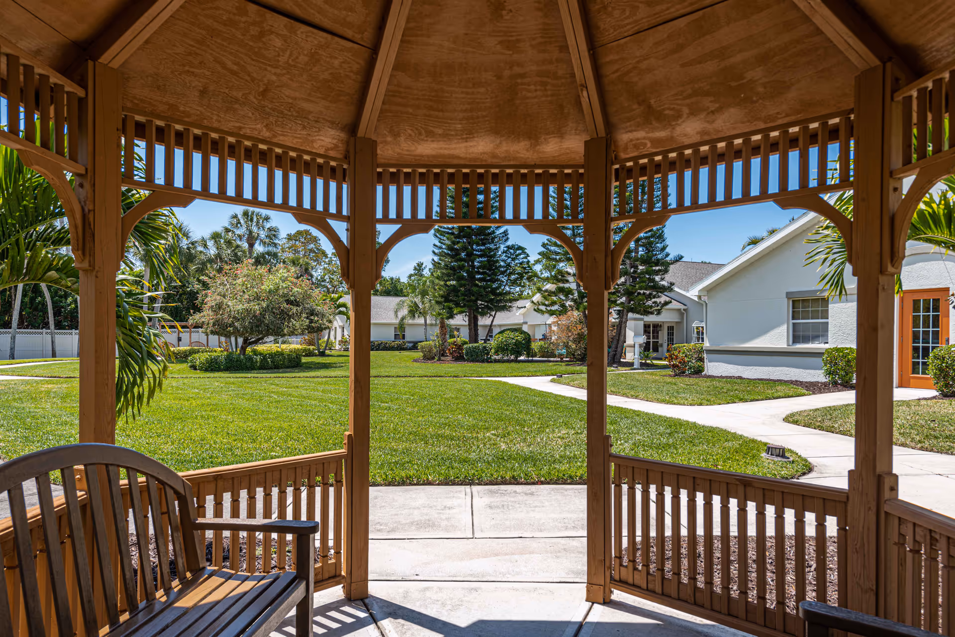 View from inside a wooden gazebo looking out onto a well-maintained grassy courtyard with palm trees, shrubs, and a paved walkway leading to white buildings with orange doors under a clear blue sky.