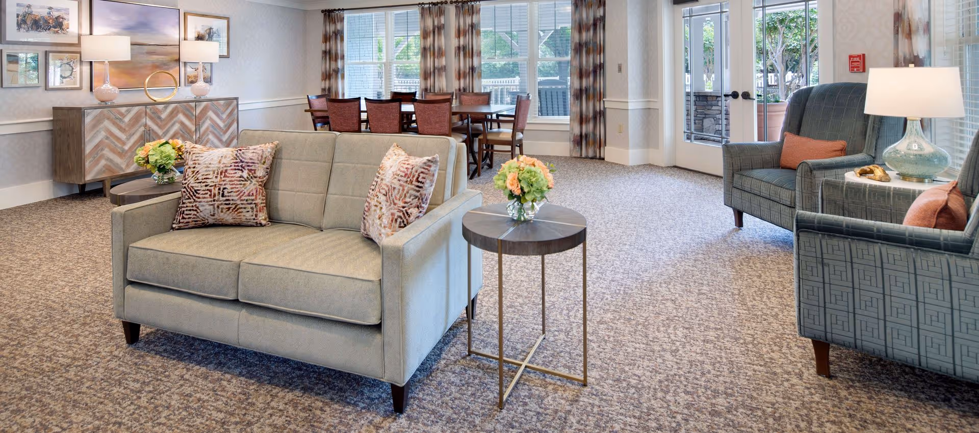 A bright and spacious living room area in a senior living facility with a beige loveseat adorned with patterned pillows, a small round side table with a floral arrangement, two blue armchairs with orange pillows, a lamp on a side table, a wooden sideboard with lamps and framed artwork above it, and a dining table with chairs near large windows and glass doors leading outside.