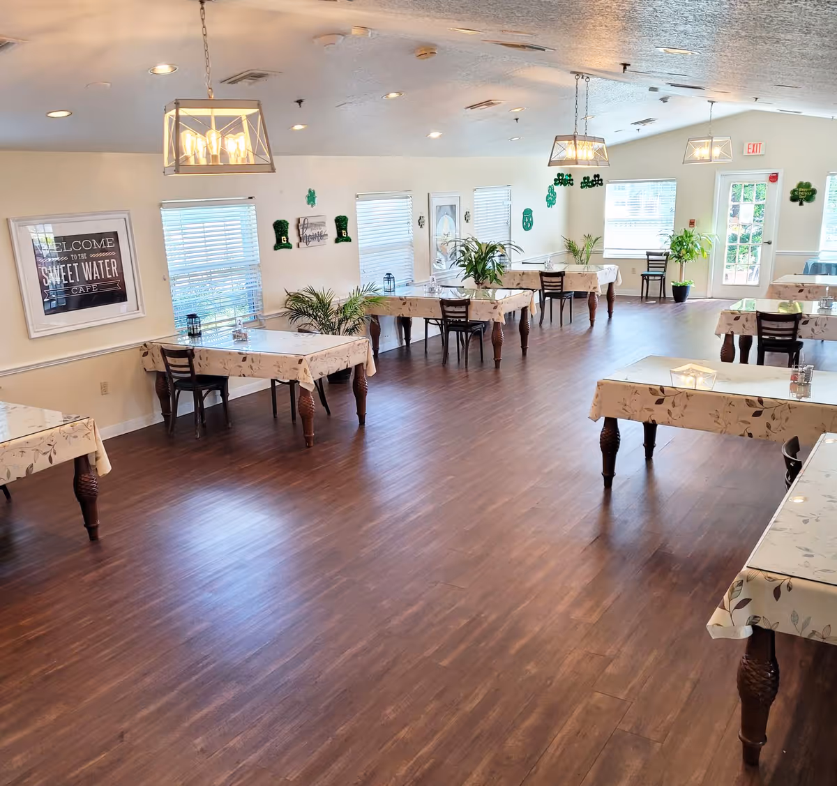 A spacious dining room with several tables covered in floral tablecloths, each accompanied by chairs. The room has wooden flooring, large windows with blinds, and hanging light fixtures. There are green plants on some tables and St. Patrick's Day decorations on the walls. A framed sign on the wall reads 'Welcome to the Sweet Water Cafe.'